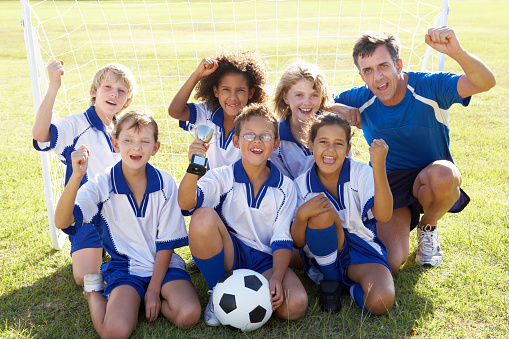 Group Of Children In Soccer Team Celebrating With Trophy By Goal On Playing Field