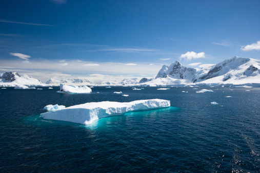A scenic view of Paradise Harbour in the Antarctic peninsula. Background of glaciers and snow topped mountains with still waters and an iceberg in the foreground