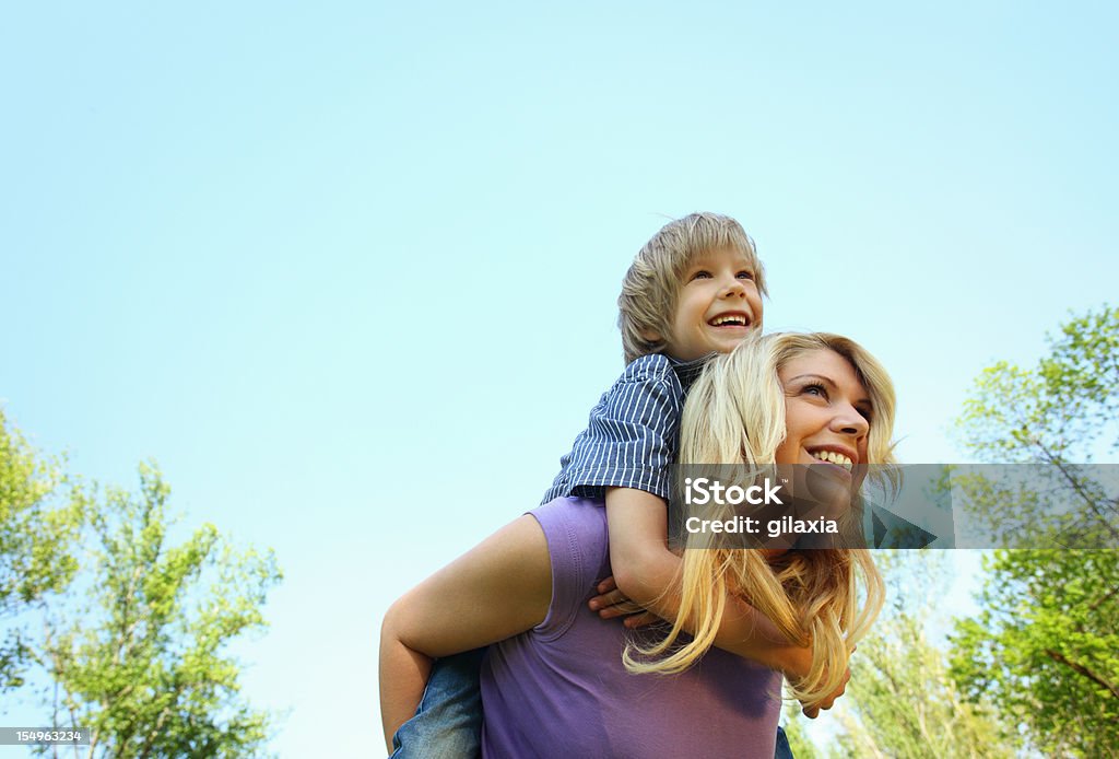 Mother and son enjoying outdoors. Mother piggybacking her son in a park. The boy is elementary age blond kid. His mother is in early 30's. Both laughing and having fun. Low angle view. Time Stock Photo
