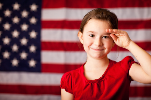 A little girl with big smile is dressed up in USA gear and ready to celebrate election day. She is smiling at the camera, wearing glasses and holding American flags. Exercise your right to vote.