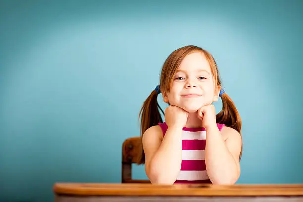 Color photo of a happy, young girl student sitting in a school desk on blue background.