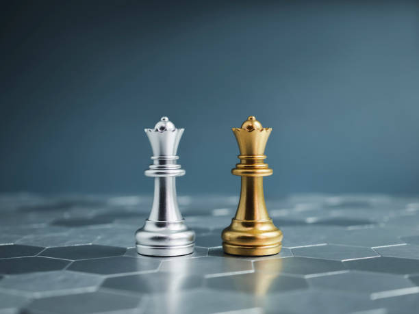Golden and silver queen luxury chess pieces standing together on a silver hexagon pattern chessboard on blue background. Leader, friend, enemy, cooperation, partnership, and business strategy concept.