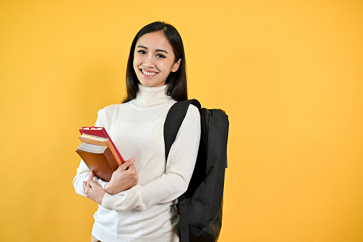 A portrait of a pretty young Asian female college student with her backpack holding her books, standing against a yellow studio isolated background. Education concept