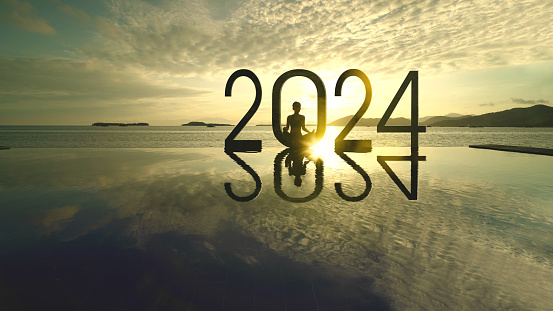 Silhouette of young woman exercising yoga while doing meditation with 2024 new year numbers on the beach at sunrise time
