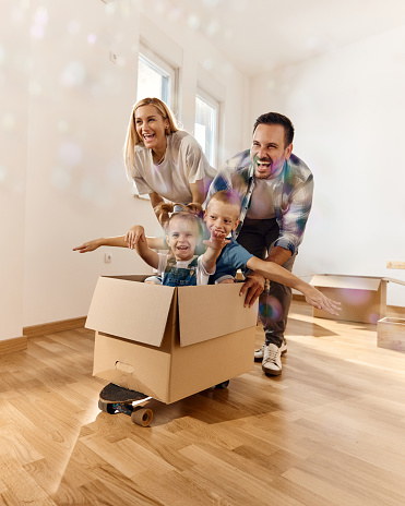 Playful parents having fun while pushing their small kids in carboard box at new apartment. Copy space.