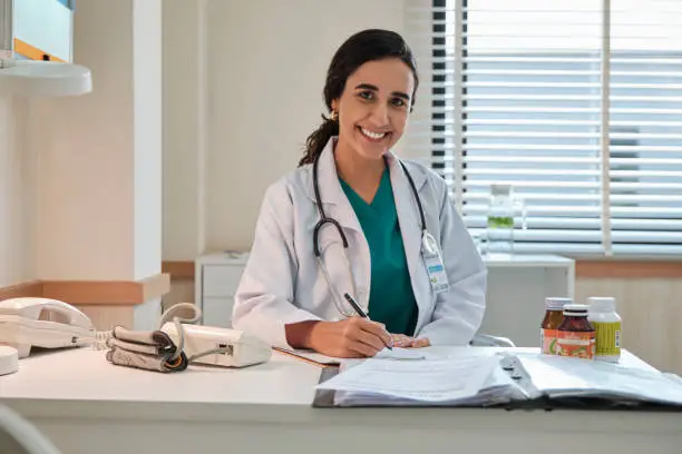 Portrait of beautiful female doctor and professional nutritionist in uniform smiling and looking at camera with supplementary foods on desk for healthy diet at hospital.