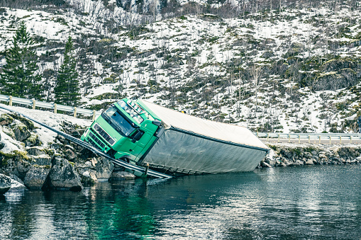 Crashed truck next to a road in a Fjord after an accident in Northern Norway. The truck has crashed through the safety barrier and driven into the cold freezing water of the Fjord next to the E10 road on the Lofoten islands in Northern Norway in winter .