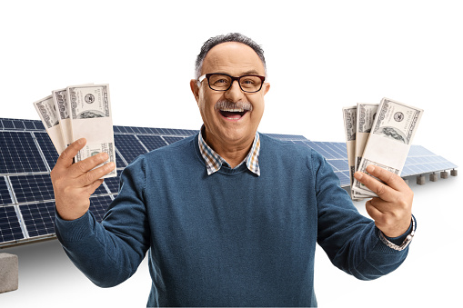 Excited mature man holding stacks of money in front of a photovoltaic farm isolated on white background