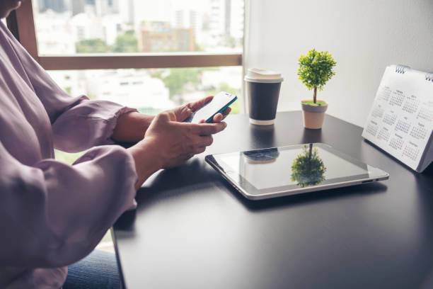 cerrar las manos de la mujer usando el teléfono inteligente comprar en línea en el escritorio de la oficina. pantalla táctil de la mujer que sostiene el teléfono inteligente abierto sitio web social. sitio web de compras en línea en computadora portá - working at home calendar laptop women fotografías e imágenes de stock