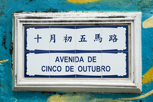 Street sign in Chinese and Portuguese made from ceramic tiles on the island of Coloane, Macau, referring to the revolution in Portugal on 5 October 1910.  The sign is on a wall which contains a mural, located on the waterfront facing the Chinese border.  This image was taken on a sunny afternoon on 5 July 2023.