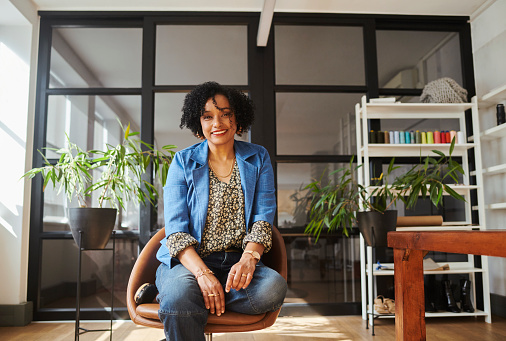 Wide shot of a creative mature businesswoman sitting with her one leg crossed on the chair while smiling into the camera. Stock photo, copy space