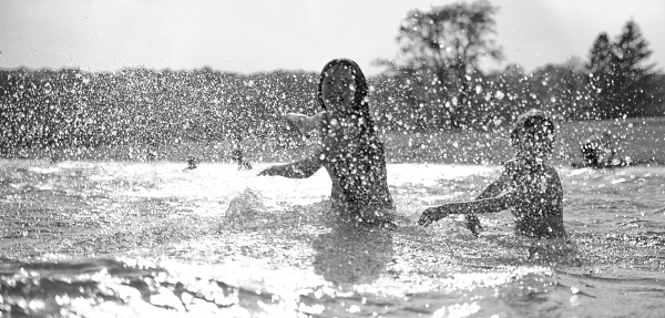 Two little girls run in water next to the beach on the lake, with lot of splashes. Black and white; backlight. 