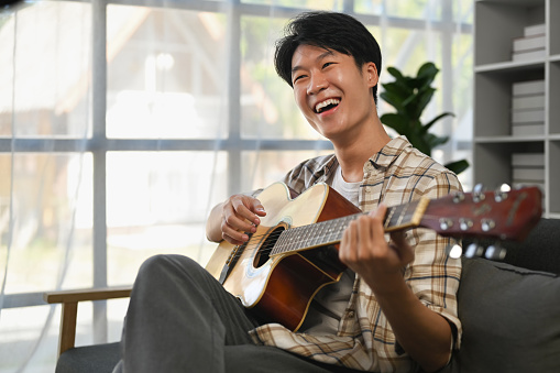 Joyful young asian man playing acoustic guitar on couch, spending leisure time in cozy living room.