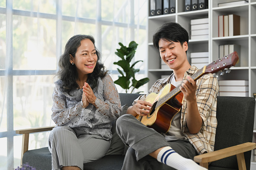 Happy family, entertainment, leisure activity concept. Young Asian man playing guitar and singing with grandmother at home.