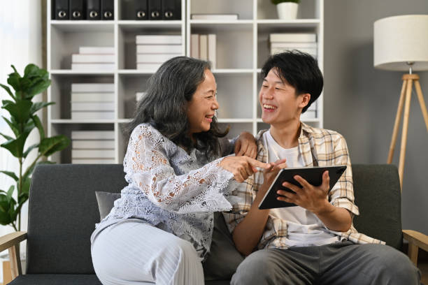 Happy young adult asian man and middle age mother sitting on sofa using digital tablet together at home.
