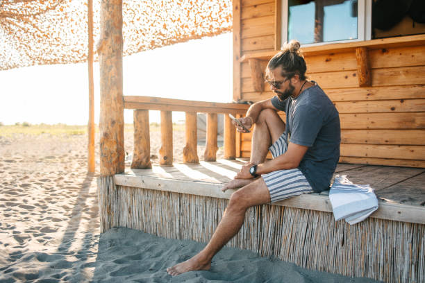 A young urban man sitting in front of a log cabin and using a phone. Wearing eyeglasses and summer casual striped clothes.
