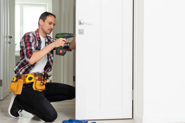 istock Maintenance Concept. Focused serious young locksmith with tattoo on hand wearing blue uniform standing on one knee in doorway installing doorknob handle, repairing wooden front door entrance 1550144099