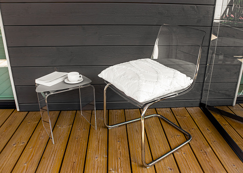 Clear transparent modern acrylic furniture in home. Chair with pillow, side table with tea cup and book. Alternative furniture material concept.