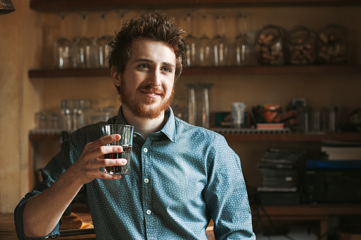 Hipster young man smiling and drinking a glass of coke with bar wooden shelves on background