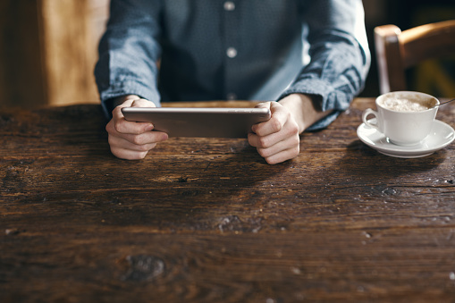 Young man having a coffee break and using a digital tablet on a rustic wooden table, unrecognizable person, hands close up