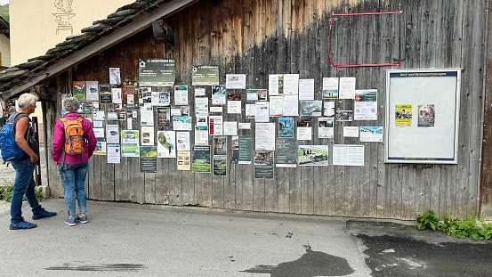 Vals, Switzerland - July, 12th - 2023: information board at a wooden wall at the village square. Two tourists reading the news.