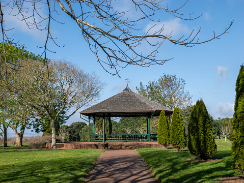 A landscape photograph of a white gazebo in a lush green park. The gazebo has benches inside to sit down and is surrounded by flowers. There is a path that can be seen cutting through the lush green grass toward the gazebo. The gazebo is surrounded in the background by large green trees.