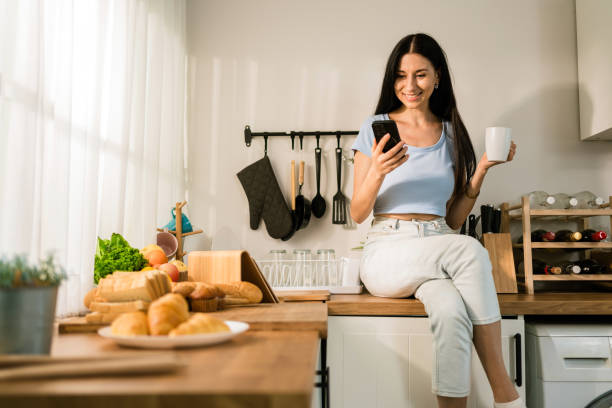 istock An attractive Caucasian woman using a smartphone while relaxing, drinking coffee and sitting on the kitchen counter at home in the morning. Happy woman shopping on mobile phone while in the kitchen. 1550296475