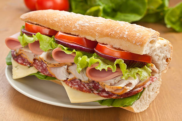 Subway Baguette Sandwich half of long tasty subway baguette sandwich with lettuce, tomatoes, ham, turkey breast, salami and cheese on a plate with ingredients at the back submarine sandwich photos stock pictures, royalty-free photos & images