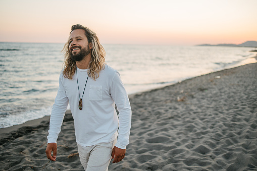 A sun tanned man with long hair in linen clothes walking by the beach at sunset. Relax