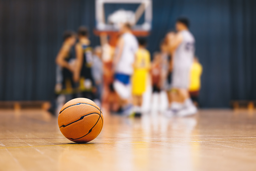 Basketball ball on wooden court. Young boys play basketball match in blurred background. Junior level sports team compete in indoor game