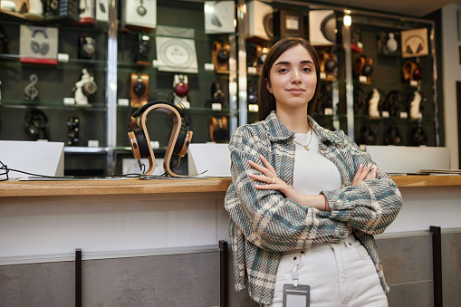 Waist up portrait of young woman working in music store and looking at camera posing confidently with arms crossed, copy space
