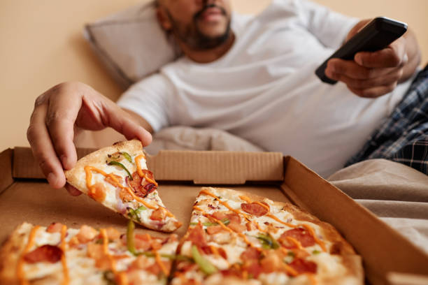 primer plano de un hombre adulto tomando una rebanada de pizza y viendo la televisión - working bed smart phone bedroom fotografías e imágenes de stock