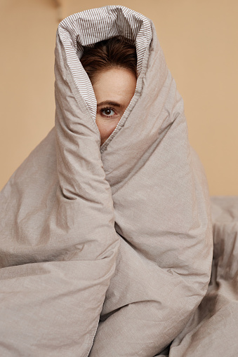 Portrait of cute young woman wrapped in cozy blanket peeking at camera with one eye