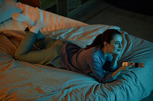 High angle portrait of adult woman watching TV in dark while lying on bed at home, copy space