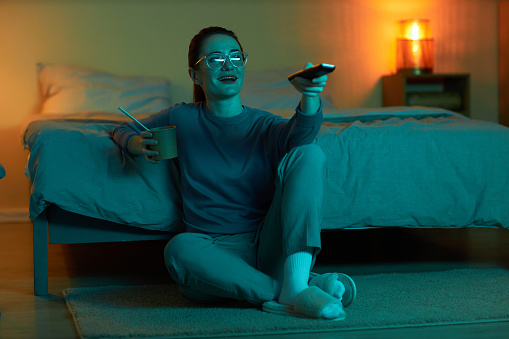 Full length portrait of smiling woman watching TV at home in dark while sitting on floor and holding remote control, copy space