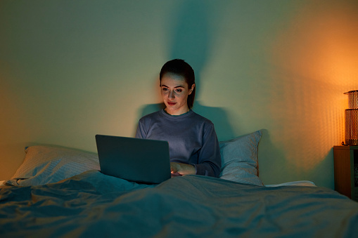 Minimal front view portrait of woman using laptop in bed at night lit by screen, copy space