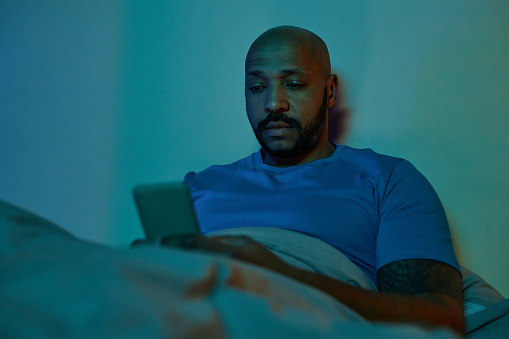 Portrait bald black man using smartphone in bed in dark suffering from insomnia