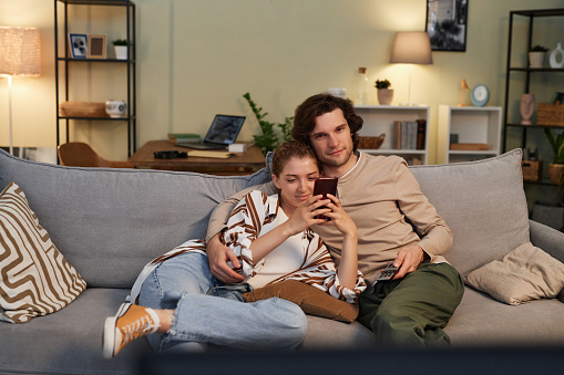 Front view portrait of young couple watching TV at home together sitting on comfortable sofa and cuddling