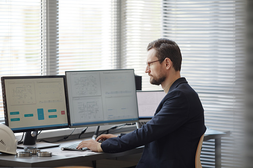Side view portrait of bearded male engineer using computer with blueprints on screen in office, copy space