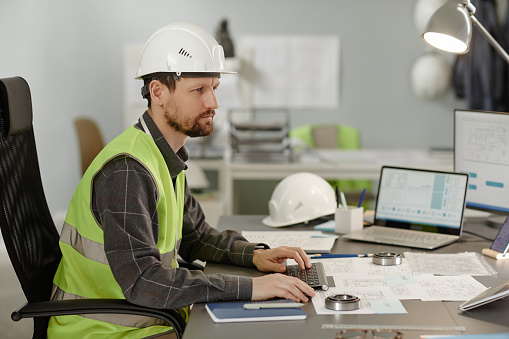 Side view portrait of bearded construction engineer wearing hardhat at workplace in office and using computer