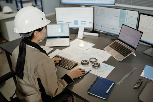 High angle portrait of female engineer wearing hardhat at workplace in office and using multiple computers, copy space