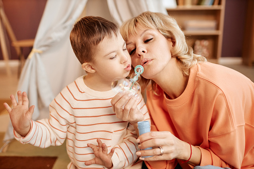 Portrait of mother and son with down syndrome blowing bubbles together while playing at home