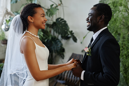 Side view portrait of young black couple as bride and groom looking at each other and holding hands