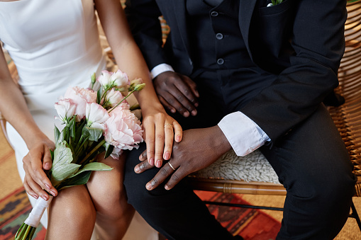 Closeup of African American bride and groom sitting on bench together and holding hands with focus on wedding rings, copy space