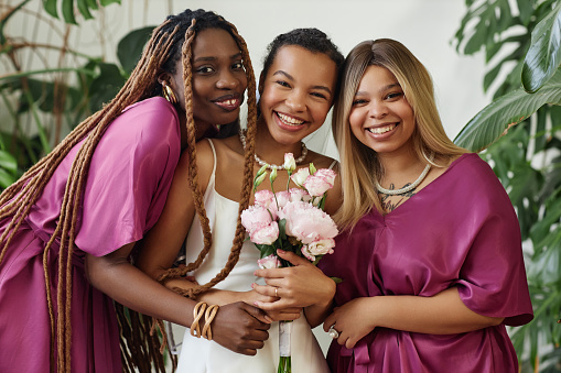 Waist up portrait of happy black woman as young bride posing with bridesmaids during wedding ceremony and looking at camera