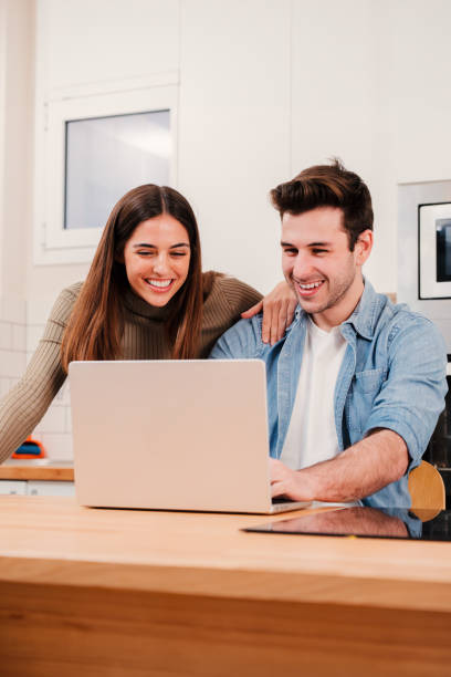 istock Vertical portrait of young couple browsing on internet at home kitchen using a laptop. Happy husband and wife planning the vacations with a computer talking and smiling together, watching funny videos 1550491280