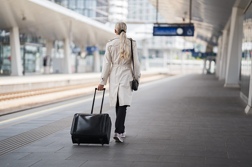 one mid adult woman with long hair pigtail wearing black trousers and beige trench coat walking along modern urban railway station platform with her trolley arriving or departing from business trip, rear view, full length