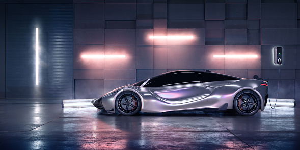 Side view of a generic silver electric sports car parked in a warehouse close to an architectural concrete wall and connected to an electric vehicle charger. Lighting Is from various fluorescent tubes on walls and ground.