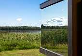 Detail view of modern mirror reflective window on building in the middle of wild nature. Luxury cottage concept. Lake, reed, trees, blue sky.