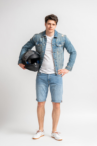 Young dark haired man in casual denim jacket and shorts, holding black protective motorcycle helmet, posing full length in studio on white background
